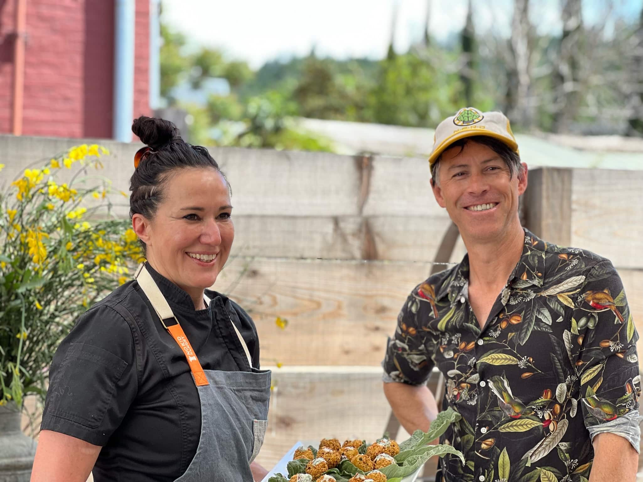 Winemaker Brook Bannister smiling in floral shirt and hat standing next to Domenica Catelli holding her delicious food.